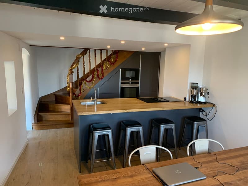 Modern kitchen area with a wooden bar counter, four metal stools, a wooden dining table with a laptop, a coffee machine, an oven, and a sink with a faucet. A staircase with wooden railings is visible in the background.