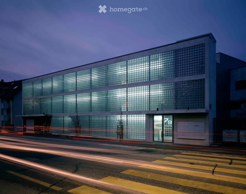 Modern two-story building with large glass windows and a clean, minimalist design, illuminated at dusk with visible light trails from passing vehicles.