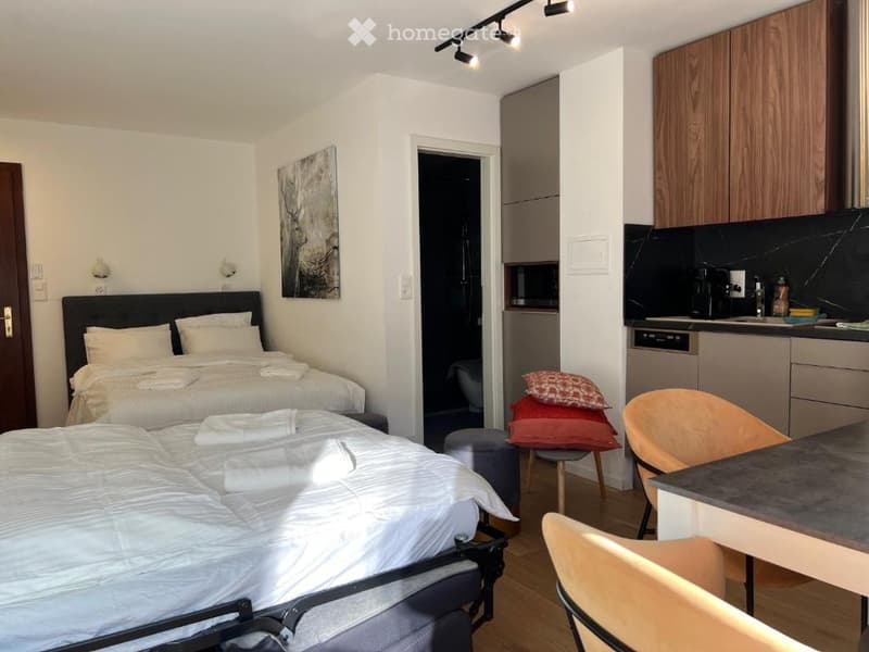 A well-lit, modern studio apartment with two single beds, a small kitchen area with cabinets and a dishwasher, a table with chairs, and a stool. The room features a painting on the wall and track lighting.