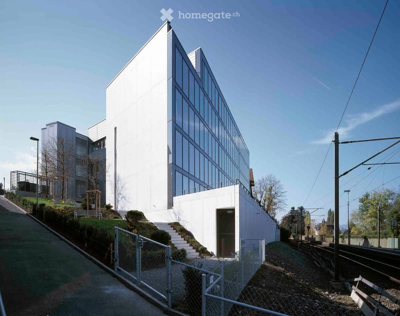 Modern white building with glass facade, raised ground floor, and steps leading up to entrance.