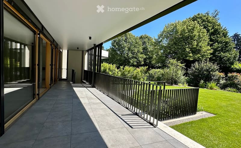 Modern balcony with tiled flooring, sleek metal railing, and large glass doors leading to the interior. The view includes a well-maintained lawn and lush greenery.