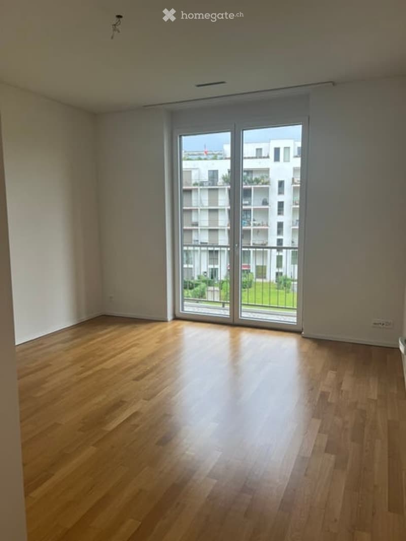 Empty room with wooden flooring and large sliding glass doors leading to a balcony with a view of a modern apartment building and greenery.