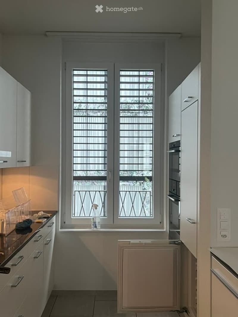 Modern kitchen with white cabinets, a countertop, and a window with blinds. A radiator is placed below the window, and an oven is visible on the right side.