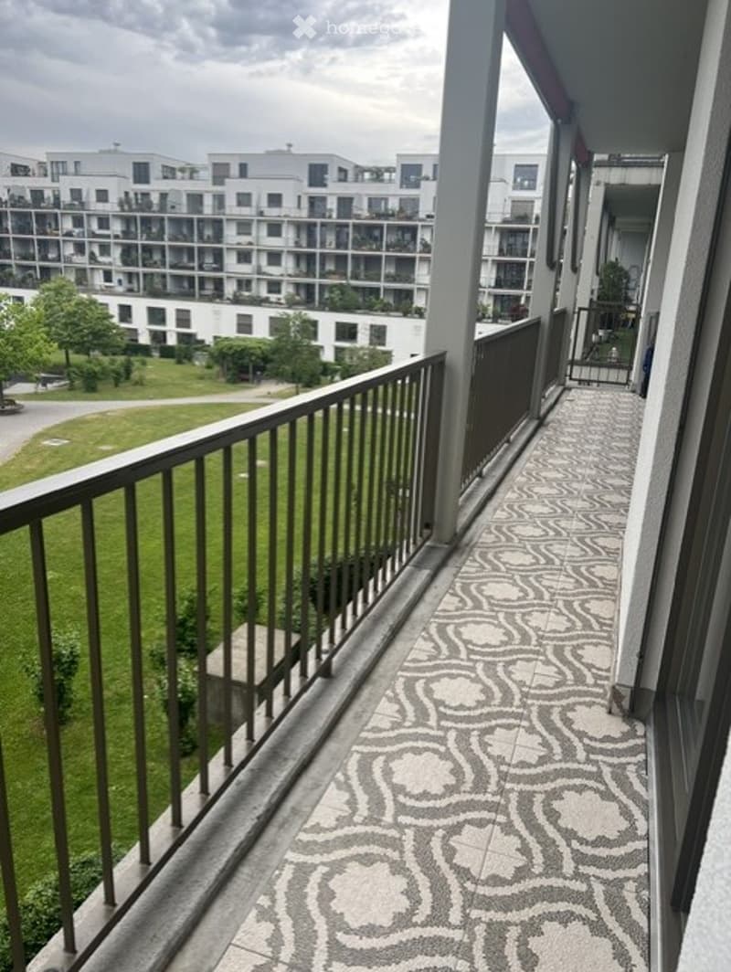Balcony with decorative tile flooring, metal railing, and a view of a green park and modern apartment buildings.