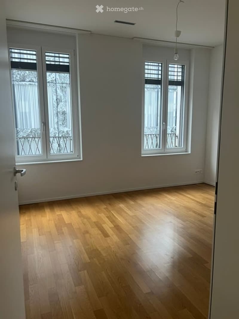 Empty room with light wooden flooring, white walls, and large windows with metal bars. No furniture or equipment present.