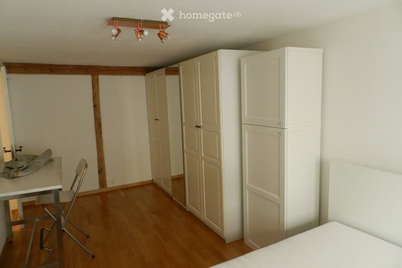 A furnished room with a bed, a chair, a table, and a wardrobe. The room features wooden flooring and white walls.