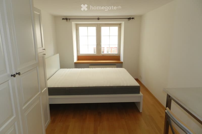 A small, furnished bedroom with a single bed, a small table, and a chair. The room features wooden flooring, white walls, and a window with a built-in wooden bench.