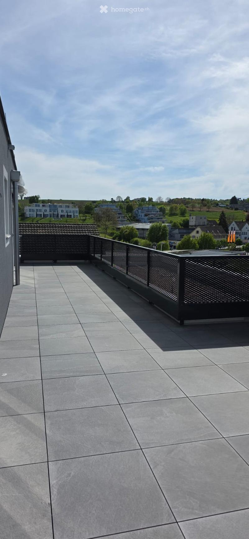 A balcony with a tiled floor and a black metal railing. The balcony offers a view of the surrounding area with buildings and trees.