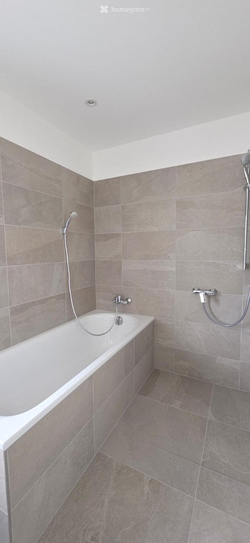 Bathroom with a bathtub, shower head, and shower hose. The walls and floor are tiled in a light gray color.