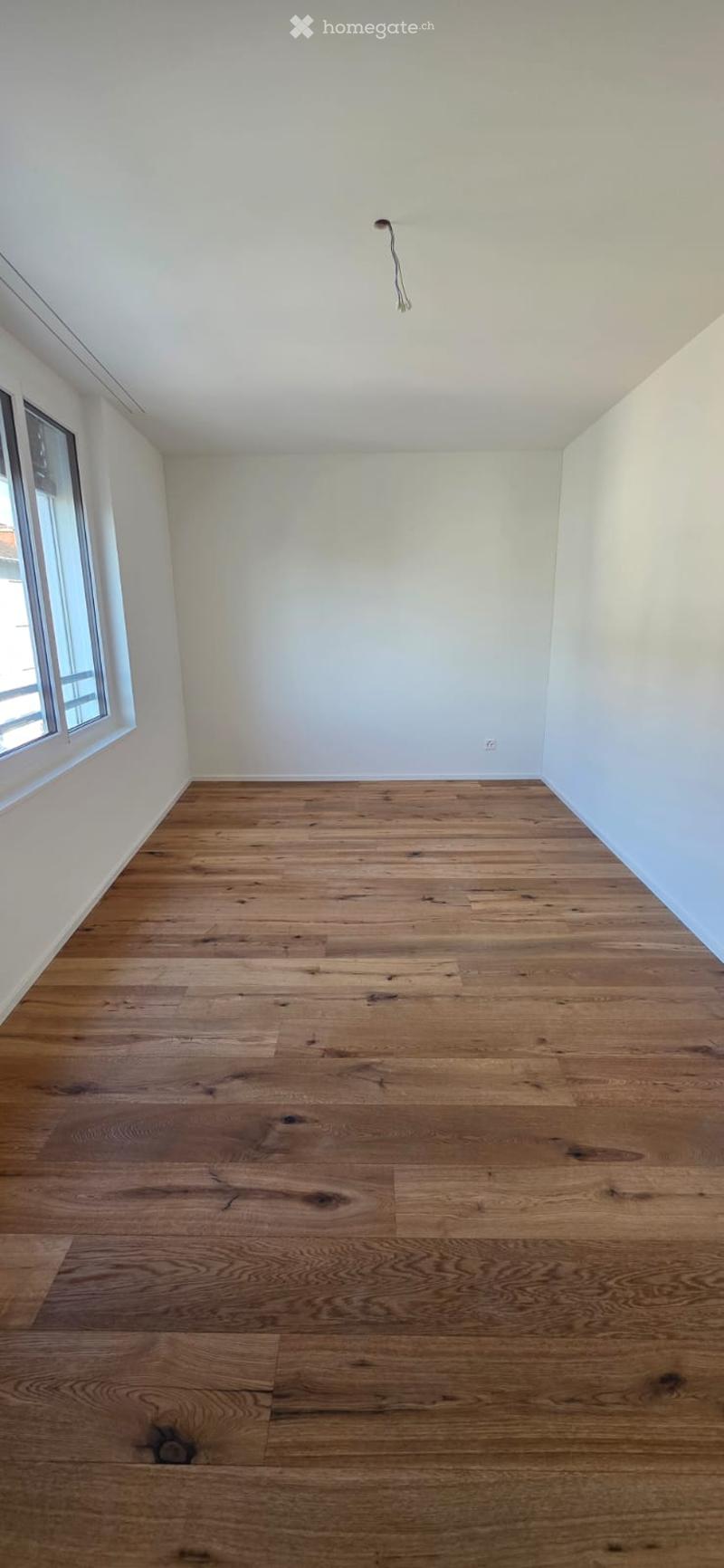 An empty room with white walls, wooden floors, and a window. No furniture or objects are present.
