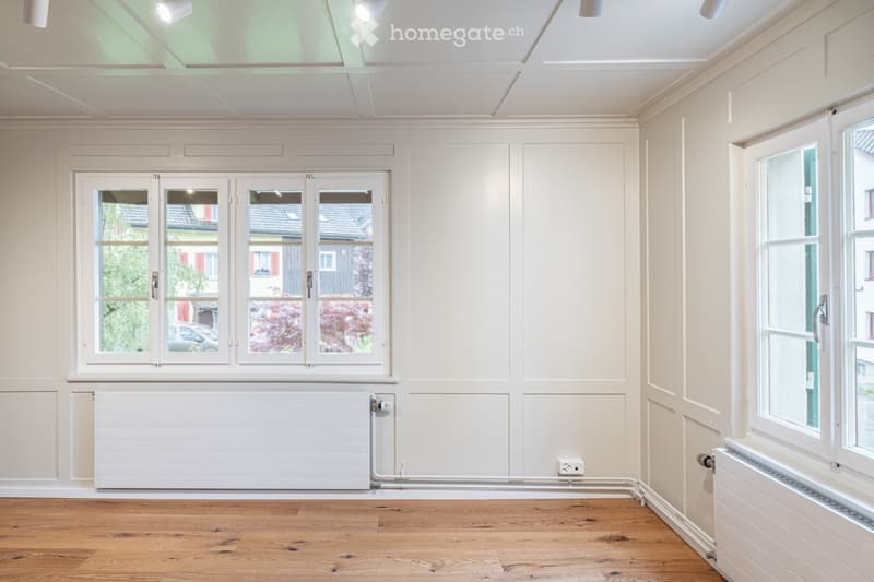 Empty room with light-colored walls, wooden flooring, large windows, and radiators. No furniture or personal items present.