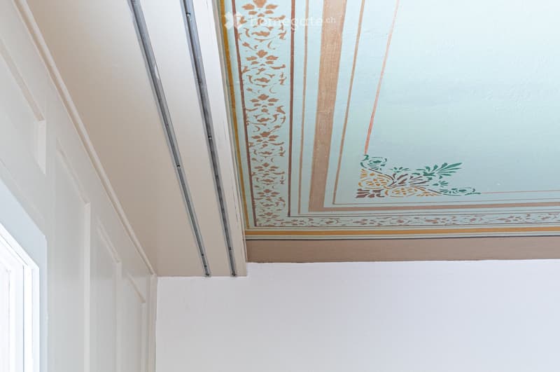 A hallway with a decorative painted ceiling featuring floral and geometric patterns in pastel colors, including light blue, gold, and green accents.