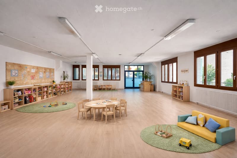 Bright, open room with wooden flooring, large windows, and modern furniture including a yellow sofa, a round table with chairs, shelves filled with toys and books, and a play area with a green rug and toys.