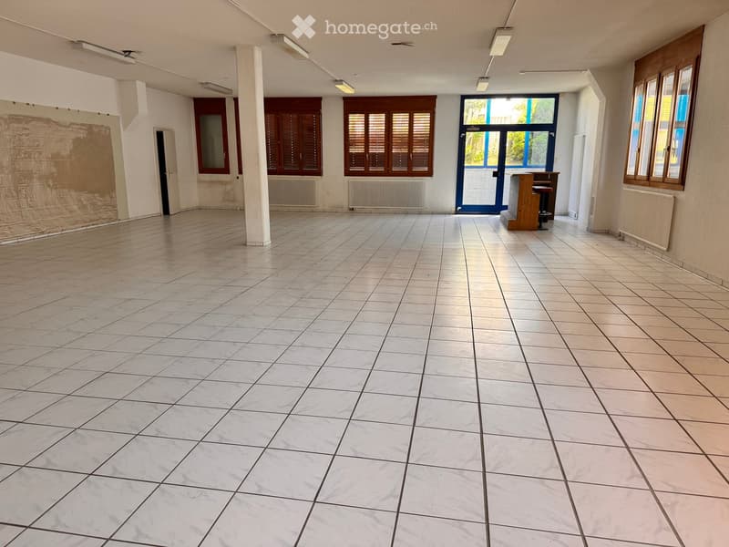 Empty room with white tiles, fluorescent lights, glass doors, windows with shutters, pillars, and radiators