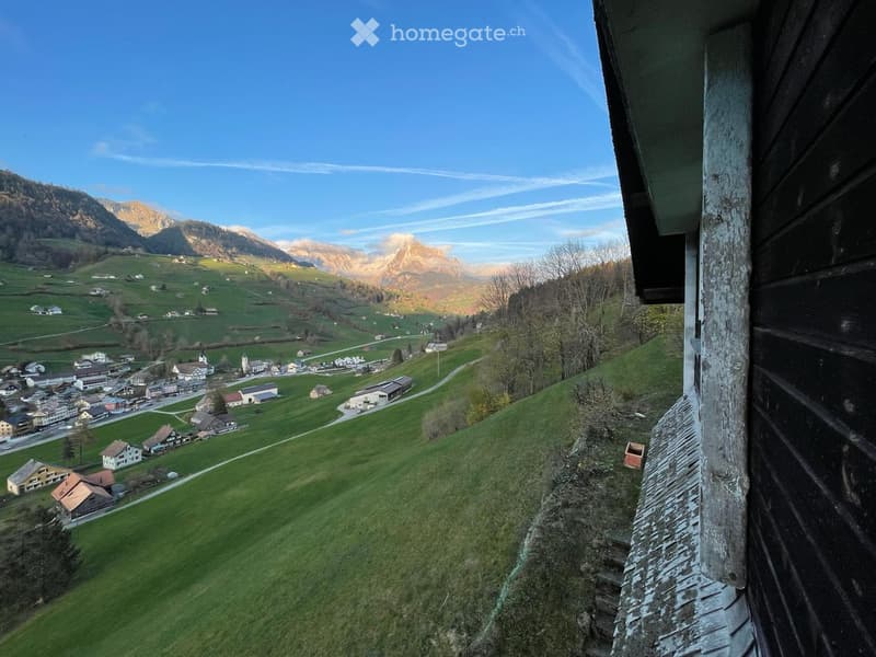 Panoramic view of a village nestled in a green valley with mountains in the background, visible from a wooden balcony or terrace.