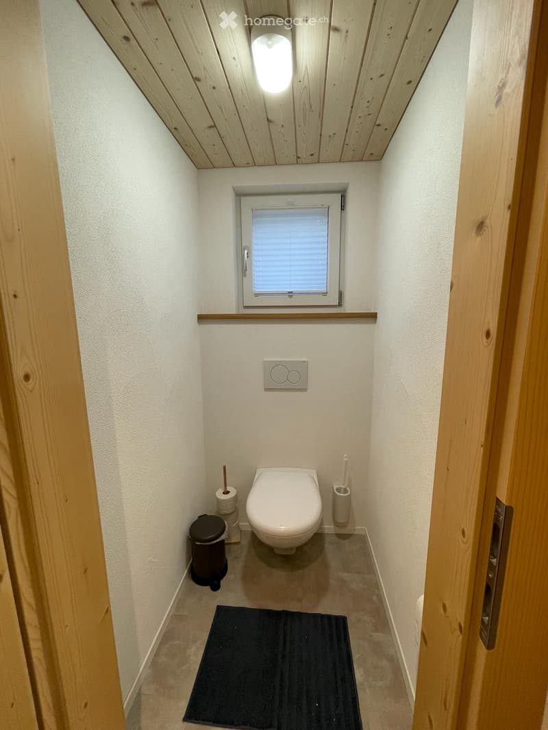 Compact bathroom with white walls, wooden ceiling, and a small window. Includes a toilet, toilet paper holders, a trash can, and a black bathroom rug.