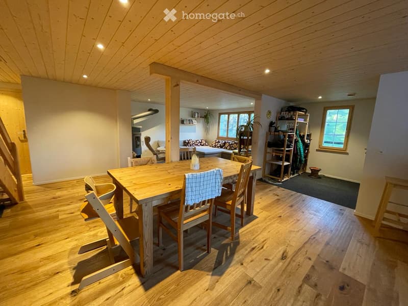 Bright, open-plan dining and living area with wooden floors, exposed wooden ceiling beams, and large windows. Includes a dining table with chairs, a sofa, and a shelf with items.
