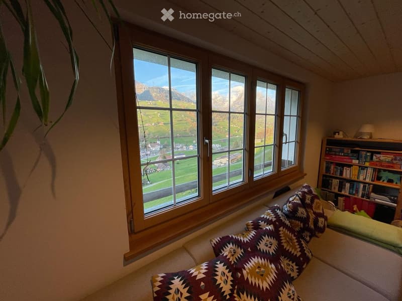 Cozy living room with large windows offering a scenic mountain view, featuring a patterned blanket on a sofa, a bookshelf, and a table lamp.