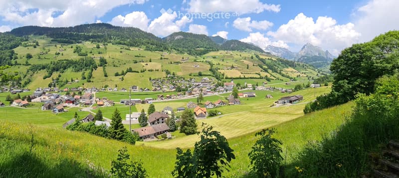 A panoramic view of a picturesque village nestled in a lush green valley surrounded by mountains. The village features scattered houses, fields, and a winding road, with a clear blue sky and scattered clouds above.