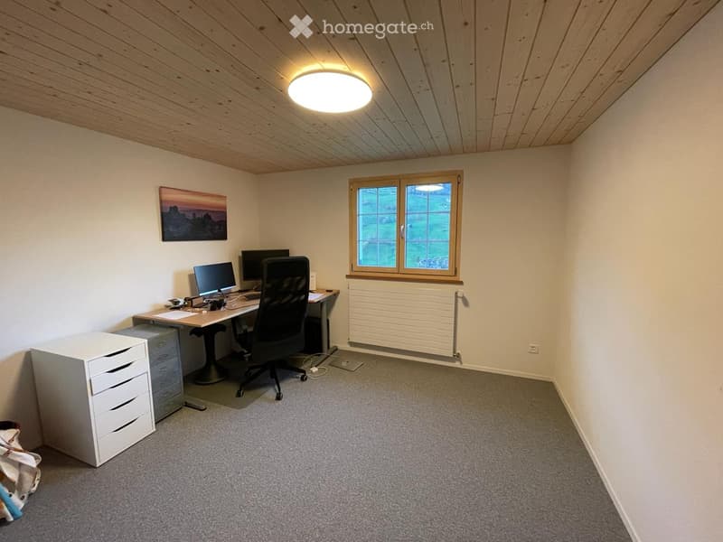 A well-lit, furnished room with a wooden ceiling, a large window, a desk with a computer and chair, a white cabinet, and a picture on the wall.