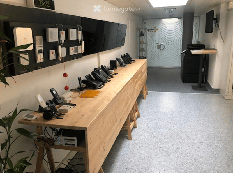 A modern reception area with a long wooden desk, multiple telephones, a large screen, and a printer. The space is clean and organized with a plant on the left side.