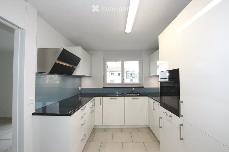 Modern kitchen with white cabinets, black countertops, and stainless steel appliances including a sink, oven, and range hood. The space is well-lit with natural light from a window.