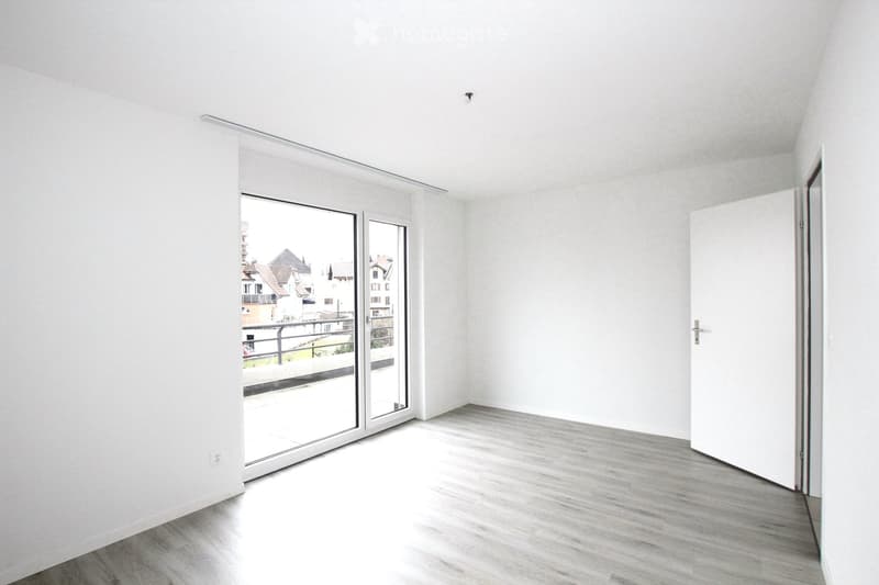 Empty room with white walls, light wooden flooring, and a large sliding glass door leading to a balcony.
