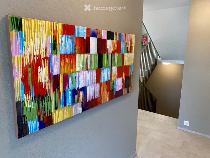 Hallway with a colorful abstract painting on one wall and a staircase leading upwards, featuring tiled flooring and neutral-colored walls.