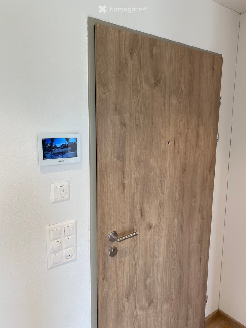 A modern hallway with a wooden door, a digital display screen, and several control panels mounted on the wall.