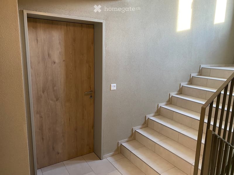 A modern hallway with a wooden door on the left and a staircase leading upwards on the right, featuring tiled flooring and light-colored walls.