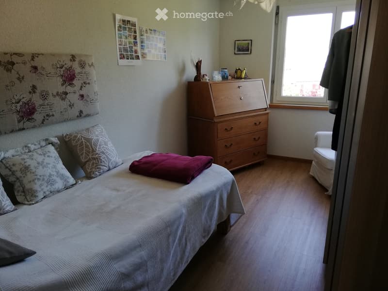 Cozy bedroom with a single bed, pillows, a purple blanket, a wooden dresser, and a small white chair near the window.