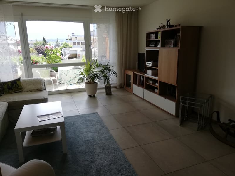 Modern living room with large windows, a sofa, coffee table, bookshelf, and a view of the balcony area.