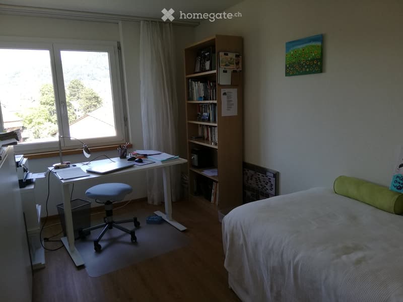 Well-lit bedroom with a bed, desk, chair, bookshelf, and a large window with curtains. The room features a modern aesthetic with a mix of functional and decorative elements.