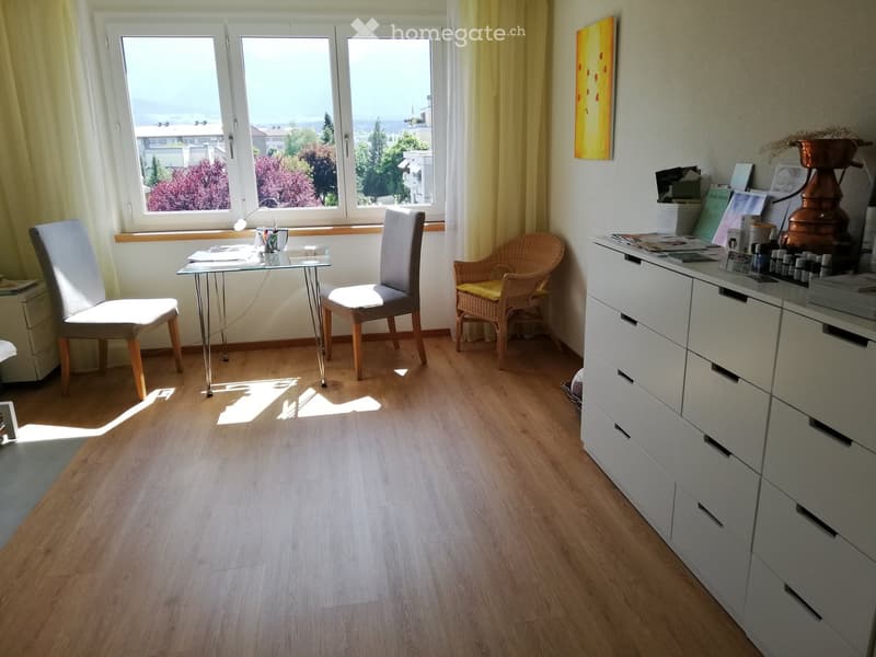 Bright room with wooden flooring, large window with yellow curtains, glass table, two chairs, and a white drawer unit with various items on top.