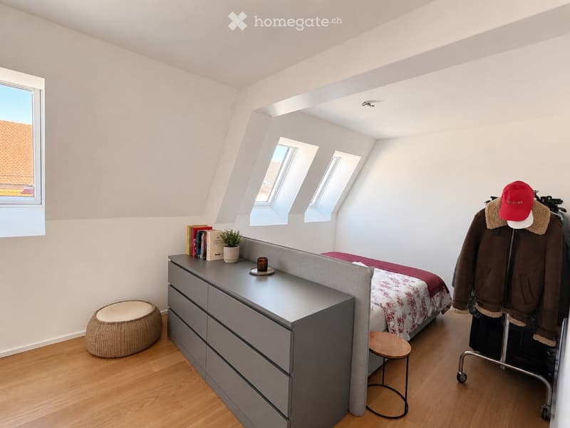 Bright attic bedroom with wooden flooring, slanted ceilings, multiple skylights, and a built-in gray nightstand with drawers. Features a bed with a patterned red and white duvet, a small round wooden stool, a woven round seat, and a coat rack with a jacke
