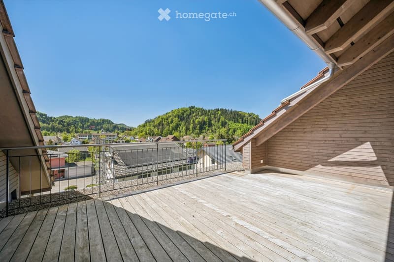 Wooden balcony with metal railing, overlooking a town with houses and green hills under a clear blue sky.
