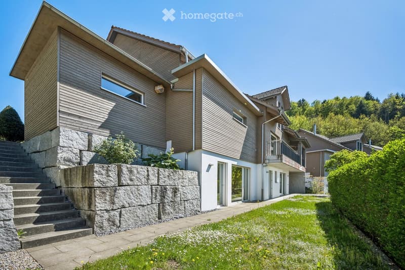Modern two-story house with wooden siding, stone steps, and a raised ground floor. Features large windows, a balcony, and a well-maintained garden.