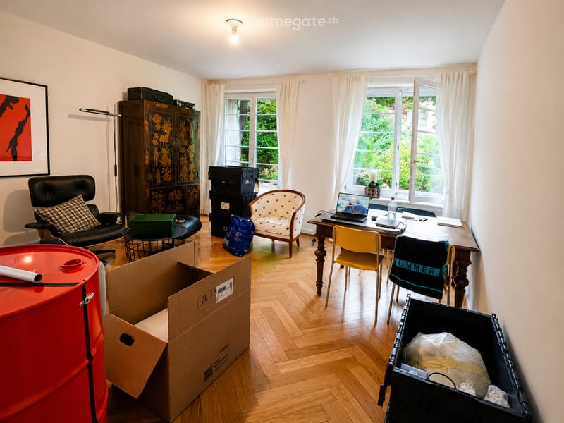 A well-lit room with wooden flooring, large windows with white curtains, a black leather armchair, a small wooden table, a laptop on a wooden desk, and several boxes and storage items.