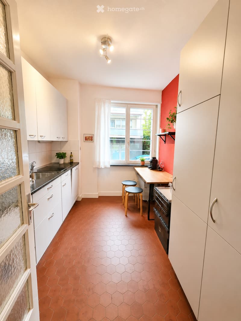 Modern kitchen with white cabinets, hexagonal terracotta tiles, a window with white curtains, a sink, a small table with two stools, and a few decorative items including a plant and a picture frame.
