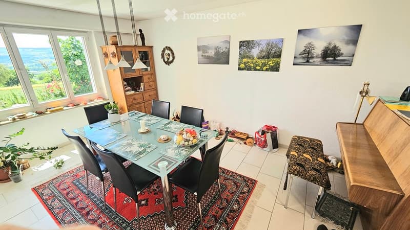 Bright dining area with a glass table, six black chairs, a wooden cabinet, pendant lights, and a view of the sea through large windows.