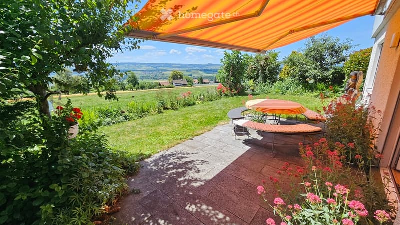 Outdoor patio with a table, chairs, and a bench, surrounded by a well-maintained garden and scenic countryside view.