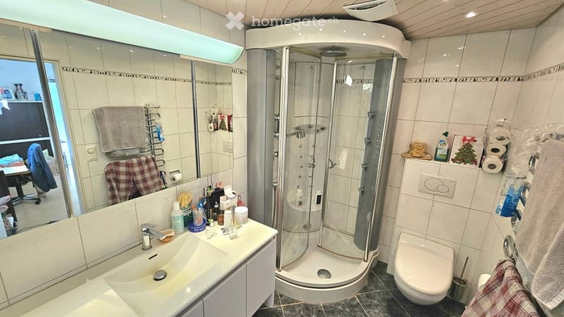 Modern bathroom with a large mirror, sink, toilet, and walk-in shower. The space features tiled walls and floor, towel racks, and various toiletries on the sink counter.