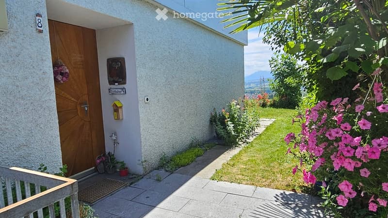 Front entrance of a house with a wooden door, decorative owl sign, and a well-maintained garden with pink flowers and a view of mountains in the background.