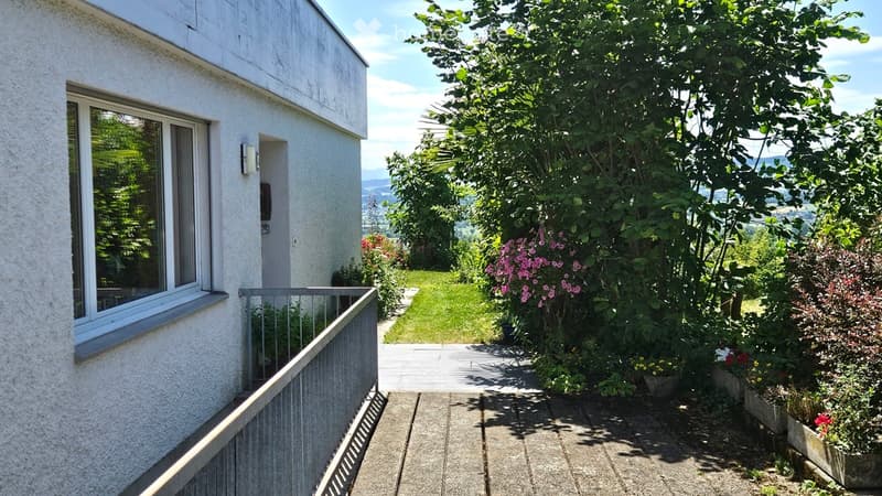 Balcony area with a view of a well-maintained garden, featuring a variety of flowering plants and trees, leading to a grassy lawn and distant landscape.
