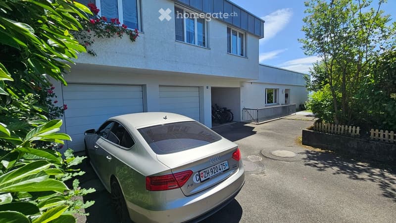 Modern two-story house with white exterior, large garage, and parking area. A silver Audi is parked in front, with bicycles stored in the garage. Greenery and a small garden are visible on the left side.