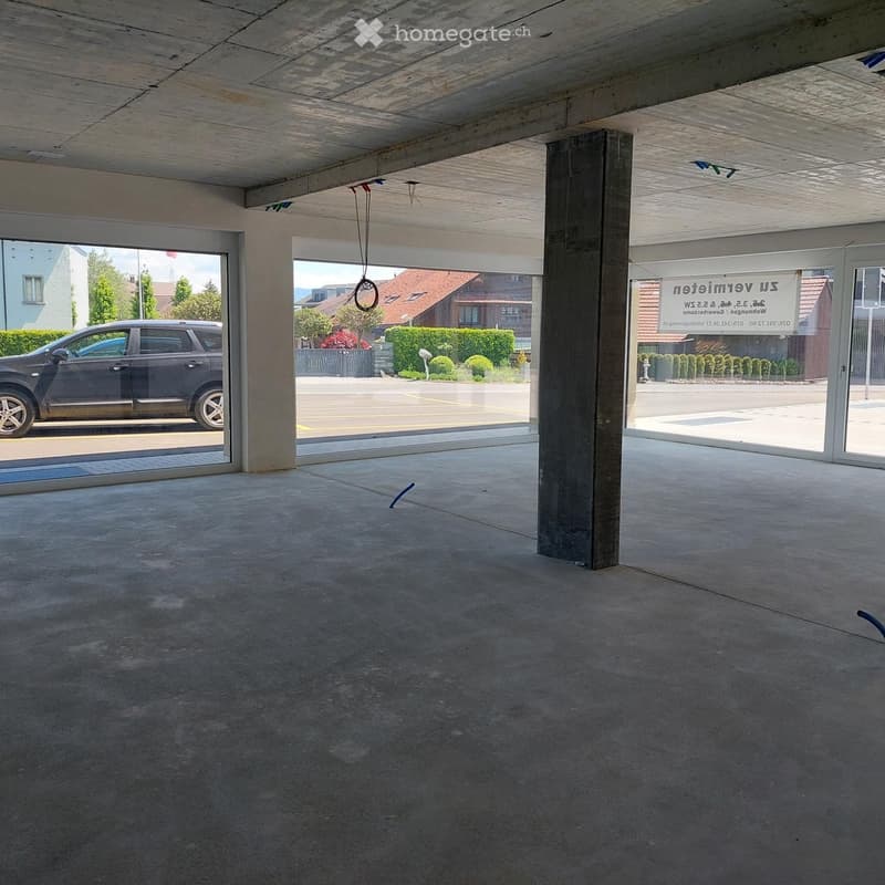 Empty concrete-floored garage with large glass doors, visible outside parking area and greenery.