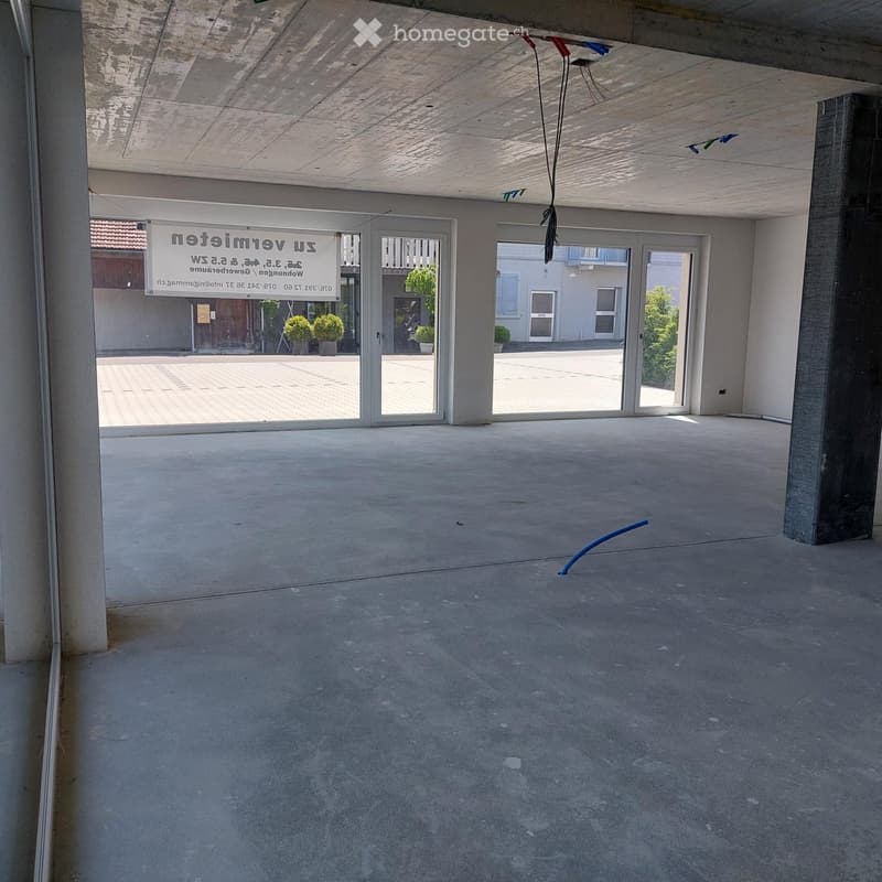 Empty room with concrete floor and walls, large glass doors leading to an outdoor area, visible signage on the wall, and a blue hose on the floor.