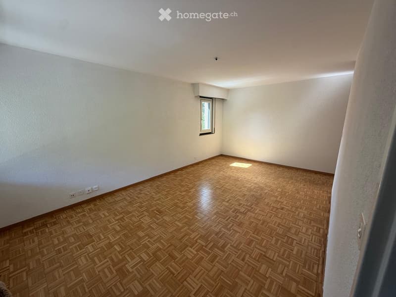 Empty room with wooden herringbone flooring, white walls, and a small window letting in natural light.