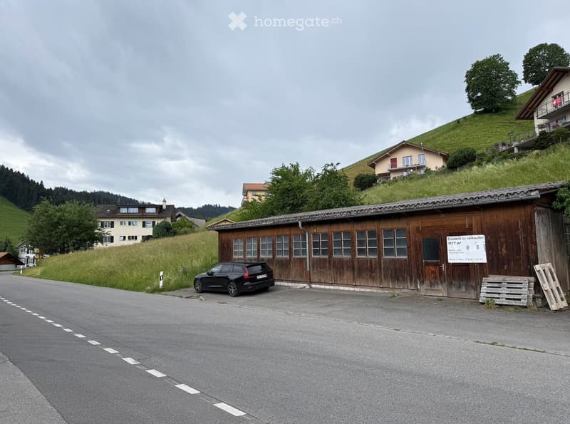 A wooden garage with multiple windows and a sign advertising a sale, located on a rural road with surrounding houses and greenery.