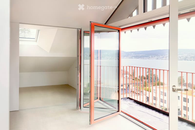 Empty room with large glass doors, skylight, balcony with railing, white walls and flooring, view of the lake and mountains